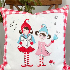 May include: A decorative Christmas pillow with a red and white gingham border. The pillow features a whimsical illustration of two carolers wearing elf hats and dresses, singing from a songbook. The pillow is on a wooden chair.