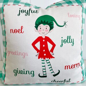 May include: A white pillow with a green and white checkered border. The pillow features a cartoon elf in a red dress and green hat. The words "joyful", "noel", "greetings", "giving", "loving", "jolly", "merry", and "cheerful" are printed on the pillow.