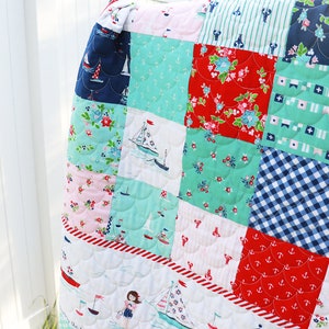 Pode incluir: Um cobertor de retalhos com tema n&aacute;utico. O cobertor &eacute; feito de v&aacute;rios tecidos em tons de azul, vermelho, branco e verde. Os tecidos apresentam padr&otilde;es de &acirc;ncoras, veleiros, listras e flores. O cobertor &eacute; acolchoado com um padr&atilde;o festonado.