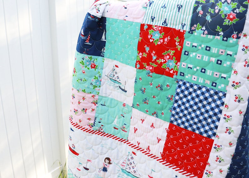 Pode incluir: Um cobertor de retalhos com tema n&aacute;utico. O cobertor &eacute; feito de v&aacute;rios tecidos em tons de azul, vermelho, branco e verde. Os tecidos apresentam padr&otilde;es de &acirc;ncoras, veleiros, listras e flores. O cobertor &eacute; acolchoado com um padr&atilde;o festonado.