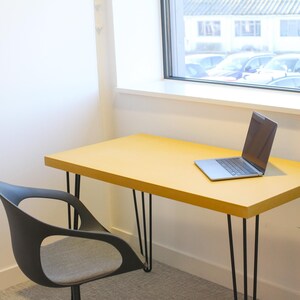 May include: A yellow desk with black hairpin legs and a laptop computer on top. A black chair with a grey seat is in the foreground.