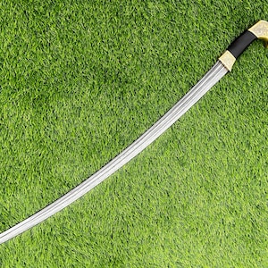May include: A curved sword with a silver blade and a black handle. The hilt is gold-colored with ornate detailing. The sword is set against a backdrop of green grass.