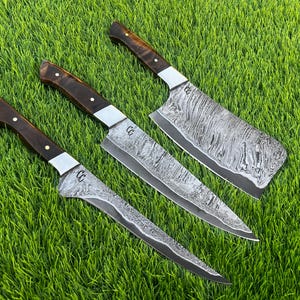 May include: Three Damascus steel kitchen knives with dark wood handles and silver bolsters are arranged on a bed of green grass. The blades have a unique, swirling pattern. The set includes a cleaver and two smaller knives.