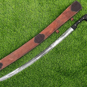 May include: A curved sword with a polished steel blade and a black handle with gold accents. The sword rests on green grass next to a brown leather sheath with dark brown accents.