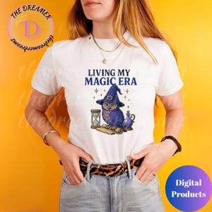 Puede incluir: Camiseta blanca con el texto "LIVING MY MAGIC ERA" y un gráfico de un búho morado con un sombrero de bruja. El diseño incluye un reloj de arena, un pergamino y una botella de poción. Producto digital.