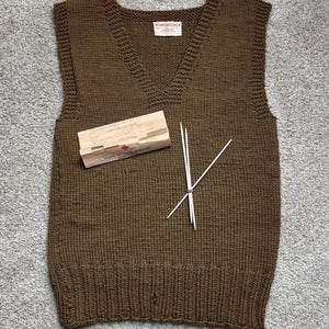 Könnte beinhalten: Eine braune Strickweste mit V-Ausschnitt. Eine kleine Holzbox mit rotem Kreuz und Stricknadeln liegen auf der Weste. Die Weste ist aus dickem, strukturiertem Garn gefertigt.