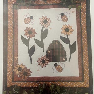 May include: A fabric quilt pattern with the text "BUSY AS BEES". The quilt design features appliqued sunflowers, bees, and a beehive. The quilt has a brown and tan border and is from Homestead Station.