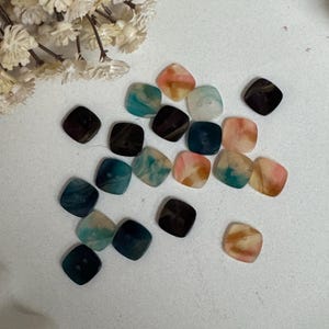 May include: A selection of square buttons in shades of brown, teal, pink, and white. The buttons have a marbled effect and are spread out on a white surface. Dried flowers are in the background.