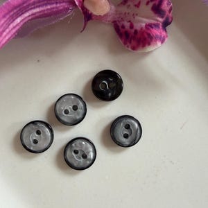 May include: Five round buttons arranged in a cluster. One button is solid black, while the other four feature a translucent, marbled grey and white pattern with a black outer rim. Each button has two central holes for attachment to fabric.
