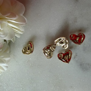 May include: Several heart-shaped charms in gold and red hues are displayed on a white marble surface. A large white flower is visible in the upper left corner. The charms have a textured, metallic finish.