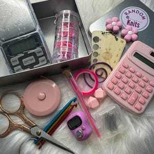 May include: A selection of knitting and crochet accessories, including a digital scale, pink calculator, scissors, measuring tape, stitch markers, and a tally counter. The kit includes a tin with the text "Random Knits" and pink flower embellishments.