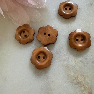 May include: Four small, light brown wooden buttons shaped like six-petalled flowers. Each button features a smooth, polished finish and two central holes for attachment. The buttons are arranged in a scattered pattern, showcasing their uniform size and shape.