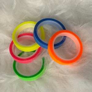 May include: Five rolls of colourful tape, including green, orange, pink, yellow, and blue. The tape is arranged in a circle on a white fluffy surface.