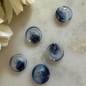 Op de afbeelding: Vijf ronde knopen met een helder, gemarmerd ontwerp in blauw- en wittinten. De knopen hebben twee gaten om te naaien en een licht gewelfde vorm. Deze knopen zijn geschikt voor diverse naaiprojecten.
