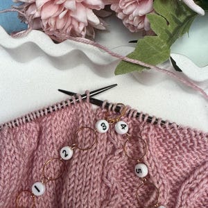 Op de afbeelding: Close-up van roze gebreide stof met zwarte breinaalden en gouden steekmarkeerders. De steekmarkeerders zijn wit met zwarte cijfers 1 tot en met 6. Roze garen en roze bloemen op de achtergrond.