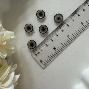 May include: Five small, round buttons with a black border and a square design in the center. A clear ruler with centimetre markings is placed next to the buttons. The buttons are on a white surface.