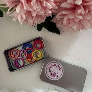 May include: A small, rectangular metal tin with a sticker that says "Random Knits". The tin is open, revealing a collection of colourful stitch markers. The stitch markers are ring-shaped and come in various shades.