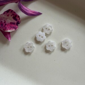 May include: Five small, white, flower-shaped buttons with two holes in the centre of each. The buttons have a smooth, slightly translucent finish and are arranged in a cluster. Each button features five rounded petals in a simple, uniform design.