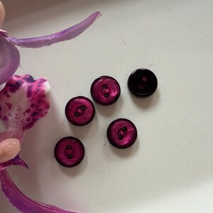 May include: Five small, round, two-hole buttons in a deep magenta and black colour palette. The buttons feature a glossy, reflective surface with a darker outer rim and a lighter, vibrant magenta centre, arranged near a purple orchid flower.