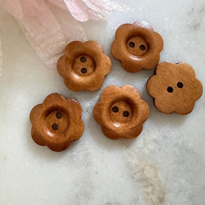 May include: Five light brown wooden buttons shaped like flowers with six rounded petals. Each button features a recessed centre with two small holes for sewing. The wood grain is visible on the smooth surface of each individual button piece.