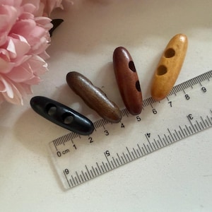 May include: Four wooden toggle buttons in shades of brown and black are arranged on a white surface next to a clear ruler. The buttons are approximately 2.5 cm to 4 cm long. Pink flowers are in the background.
