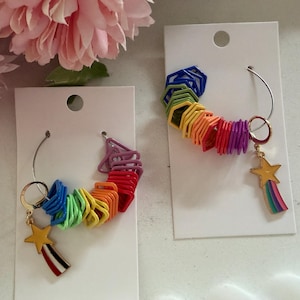 May include: Rainbow-coloured earrings with a star and rainbow charm. Each earring features a series of colourful, geometric shapes in a rainbow pattern, with a gold-coloured star charm and a rainbow tail. The earrings are displayed on white cards.