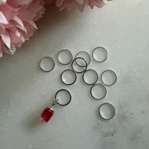 May include: A selection of silver metal rings and a red charm with a black ring. The charm is rectangular and attached to a small silver ring. Pink flowers are in the background.