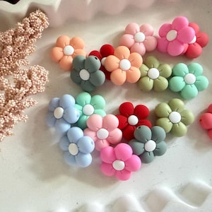 May include: A selection of flower-shaped embellishments in shades of pink, red, orange, green, and blue. Each flower has a white centre. The embellishments are displayed on a white, textured surface.