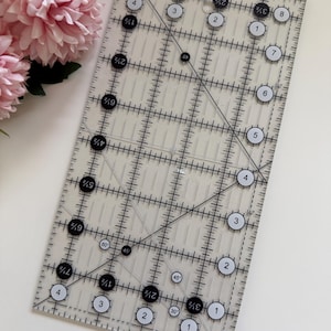May include: A transparent acrylic quilting ruler with black and white markings and measurements in inches. The ruler features a grid pattern and angle markings. Pink flowers are in the background.
