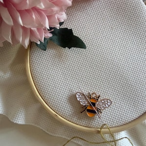 Op de afbeelding: Een borduurring met een canvas, een bijvormige bedel en een naald met draad. De bijenbedel is zwart, oranje en wit. Roze bloemen bevinden zich in de linkerbovenhoek.