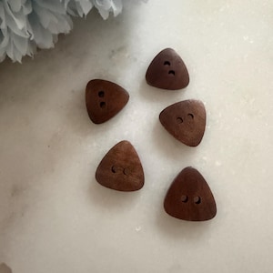 May include: Five triangular wooden buttons in a warm brown hue, each with two holes. The buttons have a smooth, polished finish and are arranged on a white surface. A light blue flower is in the background. Suitable for sewing and craft projects.