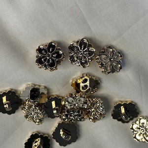 May include: A selection of flower-shaped buttons with gold-coloured rims and black, white, and silver centres. The buttons feature a detailed floral design and are arranged on a white surface.