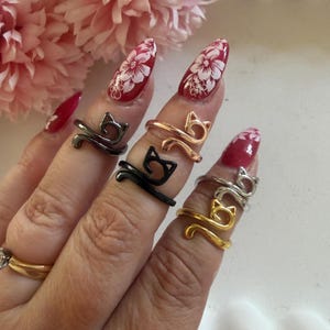 Könnte beinhalten: Eine Hand präsentiert mehrere Katzenringe in verschiedenen Metallausführungen: Schwarz, Gold, Silber und Roségold. Die Ringe werden an verschiedenen Fingern getragen, um ihr Design hervorzuheben. Der Hintergrund umfasst rosa Blumen und eine weiße Oberfläche.