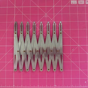 May include: A silver expanding proportional divider tool, lying flat on a pink grid cutting mat. The tool has multiple hinged arms, each connected by small rivets. The tool is designed for scaling measurements.