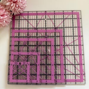 May include: Four clear quilting rulers with pink edges, black grid lines, and angle markings, nested together. The rulers are of different sizes, with the largest at the back and smallest at the front. Pink flowers are visible in the background.