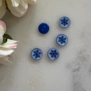 May include: Five buttons in a close-up shot. One is a solid blue button, the others are light blue with a dark blue snowflake design. The buttons are on a white surface.