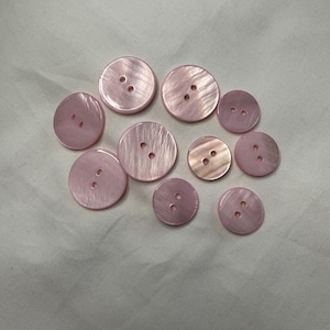 Op de afbeelding: Een verzameling ronde, roze plastic knopen met twee of vier gaten. De knopen hebben een parelmoer afwerking.