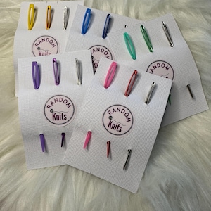 Könnte beinhalten: Sortiment an bunten Maschenmarkierern mit dem Schriftzug "Random Knits" auf den Karten. Die Maschenmarkierer sind in verschiedenen Farben, darunter lila, rosa, rot, blau, grün und silber.