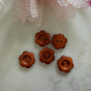 May include: Five small, brown, flower-shaped buttons with a smooth, polished finish. Each button features a rounded six-petal design and two central holes for attachment. The buttons are arranged in a cluster, showcasing their consistent warm, medium-brown wood grain colour and uniform circular shape.