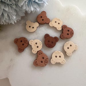 May include: A selection of wooden bear-shaped buttons in shades of brown and cream. The buttons have etched details for the eyes, nose, and ears. These craft supplies are arranged on a white surface, with a blue floral arrangement in the background.