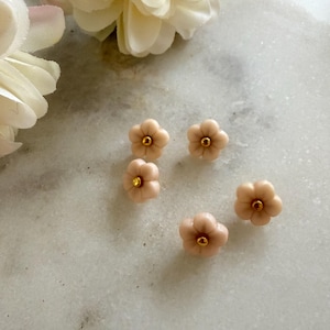 May include: Six small, pale pink flower-shaped earrings with gold centres are arranged on a marble surface. The earrings have five petals and a delicate appearance. A few white flowers are in the background.