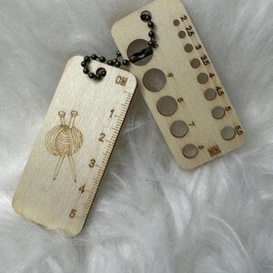 May include: A wooden knitting gauge and ruler keychain with a chain. The ruler is marked in centimetres and the gauge has nine holes for different needle sizes. The keychain features a small illustration of yarn and knitting needles.