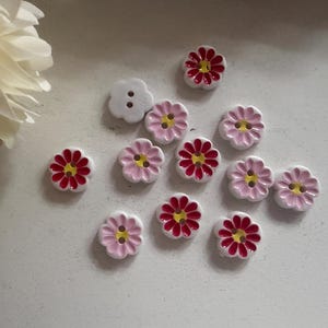 May include: A selection of flower-shaped buttons. The buttons feature a white base with pink or red petals and yellow centres. One button is a white four-petal design. These decorative buttons are suitable for sewing and craft projects.
