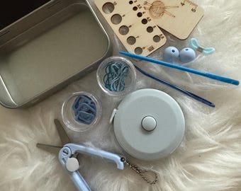 Pode incluir: Um kit de tricô azul e branco em uma lata de metal com uma régua de madeira, uma fita métrica, tesoura, marcadores de ponto e uma agulha.