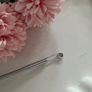 Puede incluir: Una herramienta de resorte helicoidal plateada con un extremo en bucle, probablemente para manualidades o joyería. Dos grandes flores artificiales rosas y suaves en el fondo, lo que sugiere un tema floral o decorativo.