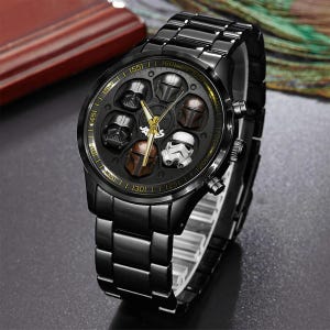 May include: Black wristwatch with a round face featuring six character helmets. The watch has a black metal band and a gold-colored second hand. The watch face includes Darth Vader, Stormtrooper, and Mandalorian helmets.