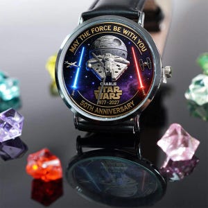 May include: A black leather watch with a silver bezel. The watch face features a Star Wars design with the text "May the Force Be With You" and "50th Anniversary". The design includes the Millennium Falcon, a Death Star, and lightsabers.