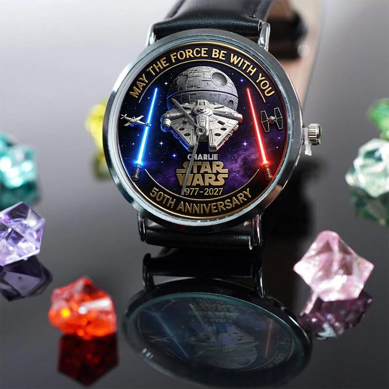 May include: A black leather watch with a silver bezel. The watch face features a Star Wars design with the text "May the Force Be With You" and "50th Anniversary". The design includes the Millennium Falcon, a Death Star, and lightsabers.
