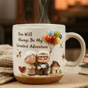 Puede incluir: Taza de cerámica color crema con base marrón, con una figura de pareja y el texto "You Will Always Be My Greatest Adventure". La taza tiene un asa y está decorada con globos, estrellas, un corazón y una casa.