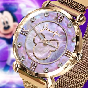Könnte beinhalten: Goldfarbene Armbanduhr mit Perlmuttzifferblatt. Die Uhr verfügt über römische Ziffern, Kristallakzente und ein von Mickey Mouse inspiriertes Design. Das Armband ist im Gold-Mesh-Stil gehalten, und das Zifferblatt trägt den Namen "Jennifer".